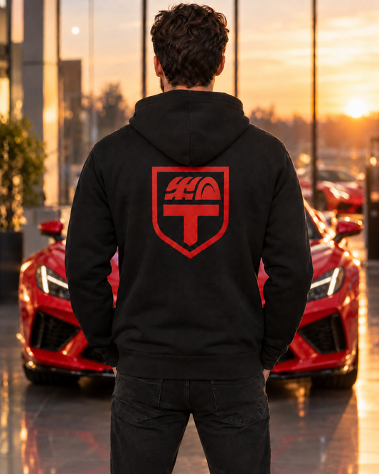 Back view of a man in a black Tread & Torque pullover hoodie with the large red shield crest across the upper back, in a Corvette showroom at sunset