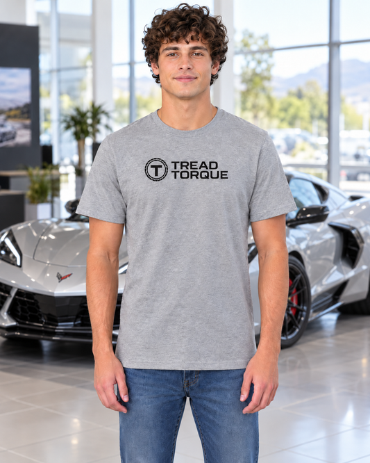 Man in a heather grey Tread & Torque classic tee with the black roundel logo on the chest, standing in a showroom beside a silver Corvette C8