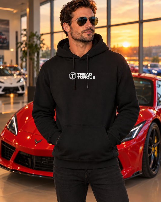 Man in a black Tread & Torque pullover hoodie with the white roundel logo on the chest, standing in a showroom beside a red Corvette at golden hour